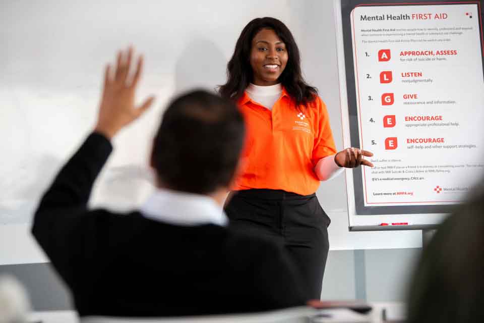 a woman in an MHFA shirt teaches at the front of a classroom