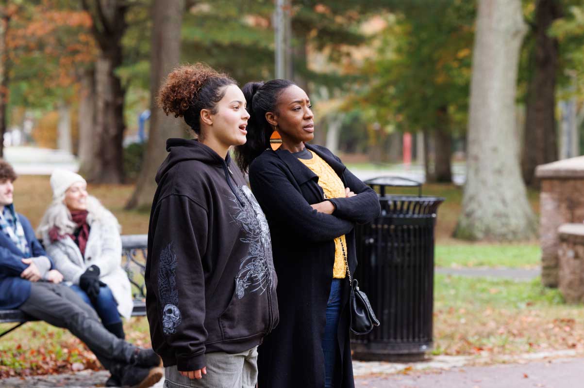 two women stand together outside in a park talking and looking at something