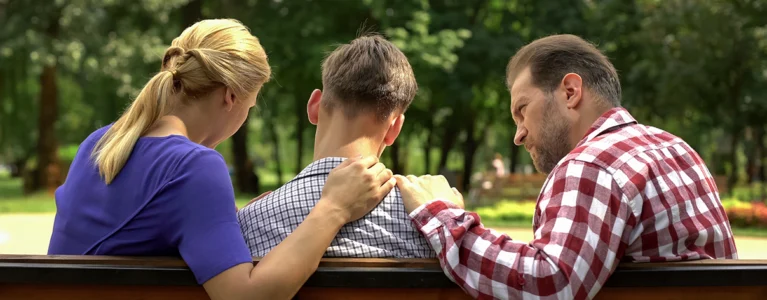 a teen and his parents sit on a park bench