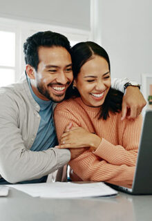 a man and woman sit together and hug in front of a laptop on a counter